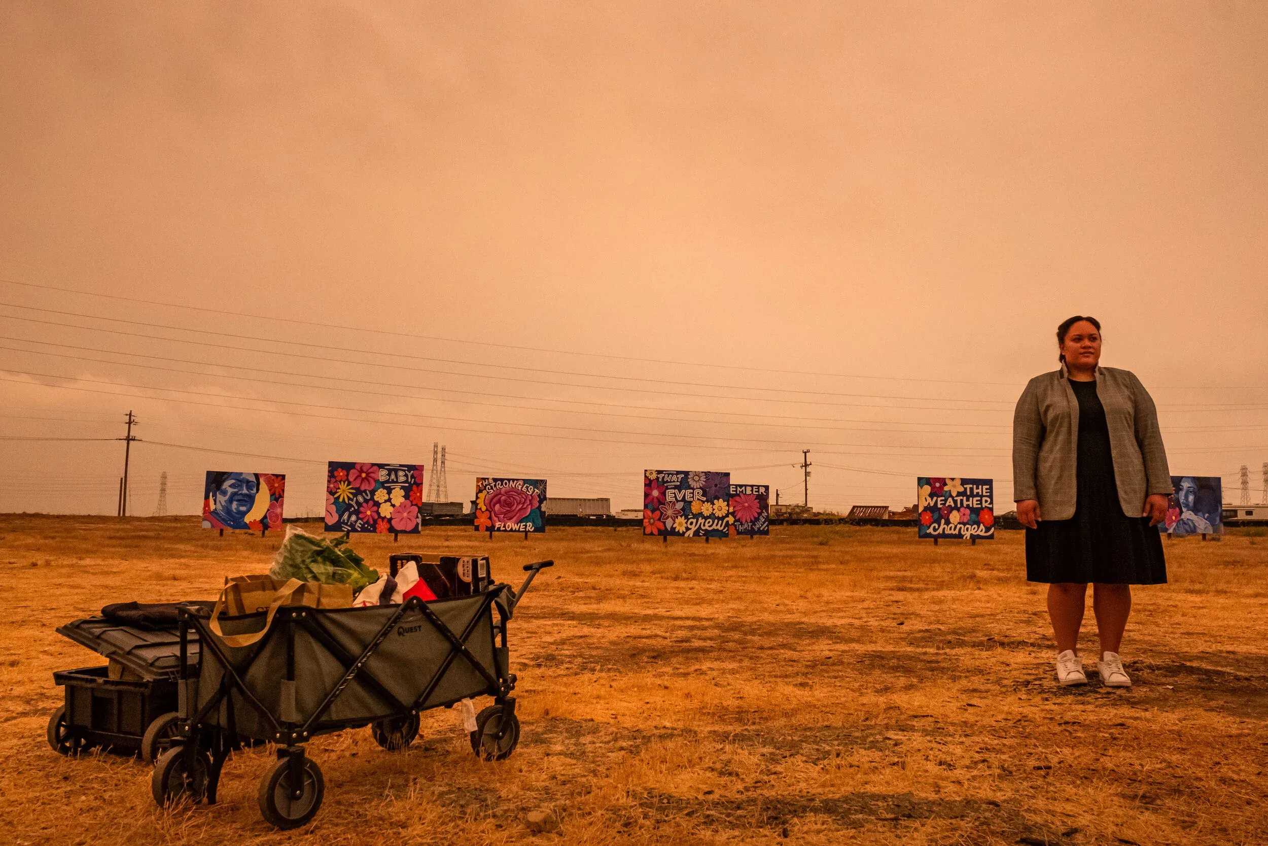 Mele Latu standing in a field with colorful murals behind her and a wagon of food supplies, under an orange wildfire sky